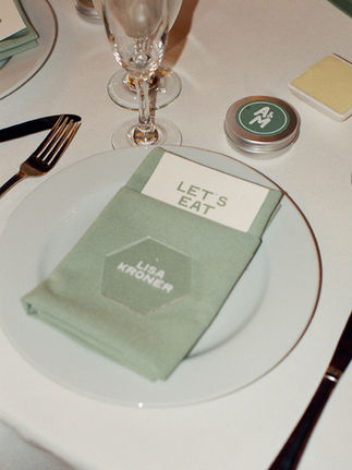A formal place setting with a green napkin neatly folded on a white plate, holding a 'LET'S EAT' card and a name card for 'Lisa Kroner.' A monogrammed tin and glassware complete the setting.