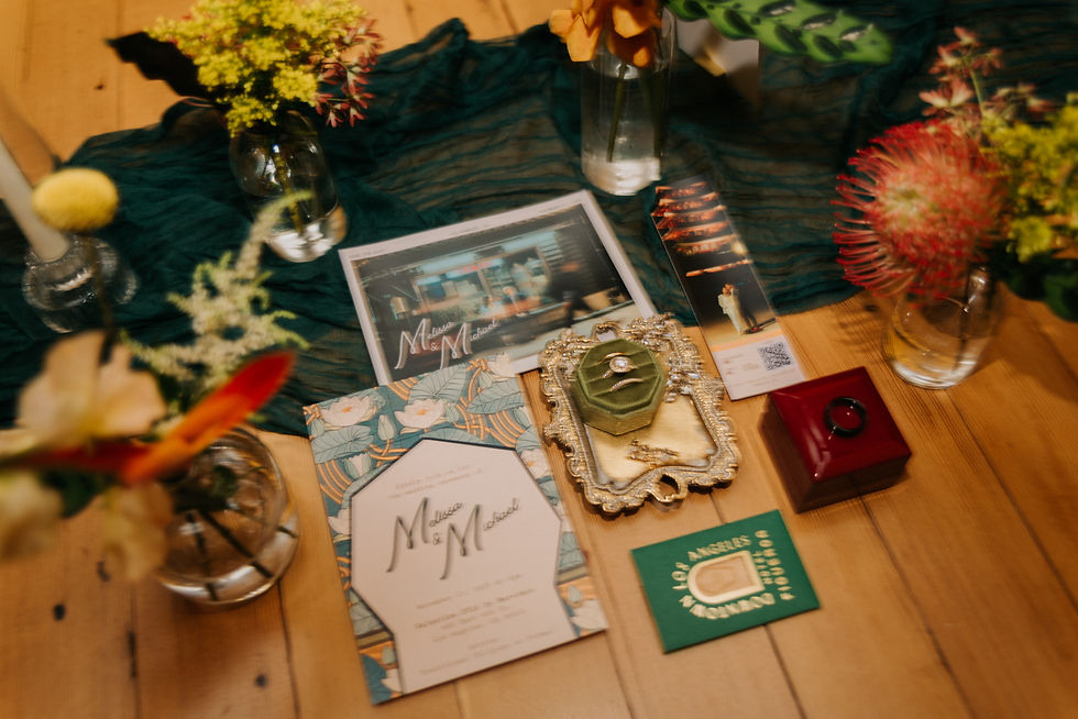 Wedding invitations with floral patterns, flowers in vases, a golden tray with a ring, and a red box on a wooden table. Text reads "Melissa & Michael".