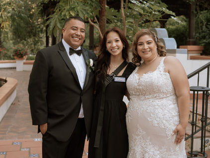 Bride in a white wedding dress, groom in a tuxedo, and Love and Life Events lead planner Dalavy smiling together in a lush garden wedding setting.
