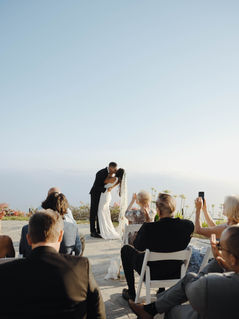 A couple walks hand-in-hand up stone steps, surrounded by palm trees and guests, set against a bright sea background. Joyful wedding scene.
