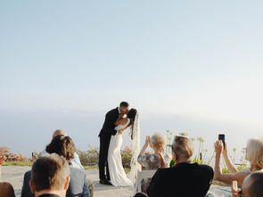 A couple walks hand-in-hand up stone steps, surrounded by palm trees and guests, set against a bright sea background. Joyful wedding scene.
