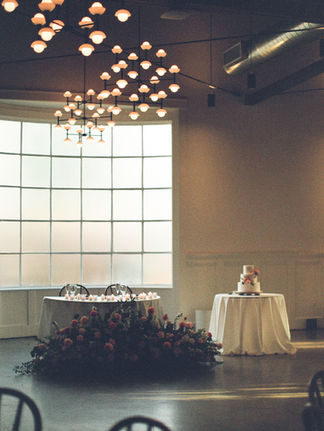 A romantic wedding reception scene with a sweetheart table in front of a large industrial-style window. The table is adorned with floral arrangements and illuminated by a ceiling full of hanging pendant lights. A cake table sits off to the right.