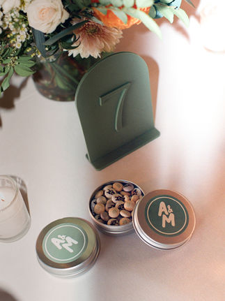 A close-up of a wedding table centerpiece featuring a green table number '7' sign, a floral arrangement, and two small tins with the initials 'A&M' on top, one open to reveal snacks.