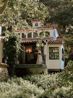A couple stands on a balcony of a white, rustic house surrounded by lush greenery. Sunlight filters through trees, creating a serene mood.