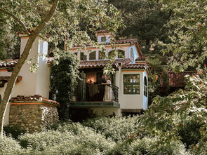 A couple stands on a balcony of a white, rustic house surrounded by lush greenery. Sunlight filters through trees, creating a serene mood.