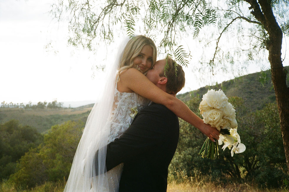 Bride smiles at the camera as the groom carries her in his arms, set against a scenic mountain and sky backdrop on their wedding day.