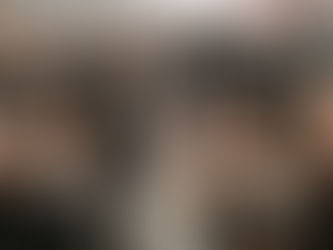 A bridal party in black dresses and suits celebrates. The couple kisses, surrounded by laughing friends holding white bouquets. Outdoor setting.