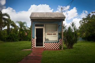 A small, blue, standalone building labeled "Charles Barber Shop" features a red-and-white checkered tile facade on its lower half and a "Closed" sign in the window, sitting centrally on a grassy lawn surrounded by green trees under a bright blue sky with fluffy white clouds.