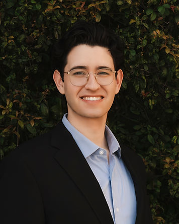 A color portrait of a man with wavy, dark brown hair and brown eyes, wearing a black suit over a blue button-down shirt. He is wearing thin-framed, gold round glasses and has a thoughtful expression. He stands centered in front of a lush, vibrant green hedge, with the natural greenery providing a clean and professional contrast to his formal attire.