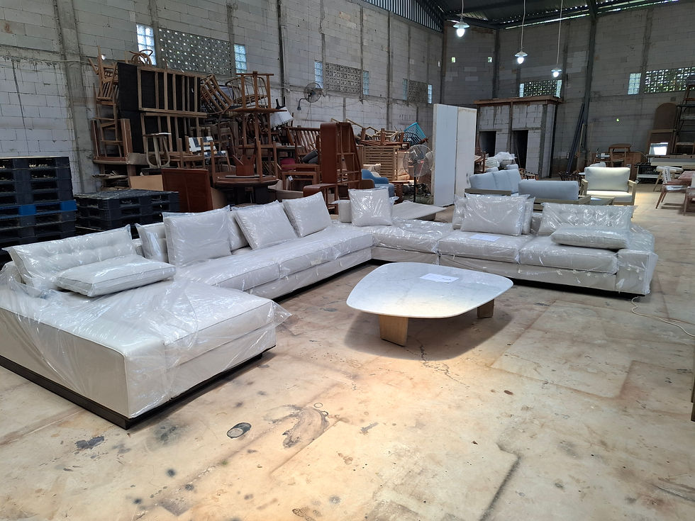 Plastic-wrapped white sectional sofas and a low table sit in a furniture warehouse, with stacked chairs and tables behind.