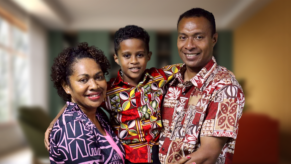 Smiling family of three in colorful patterned shirts, standing together in a cozy living room. Bright window light in the background.