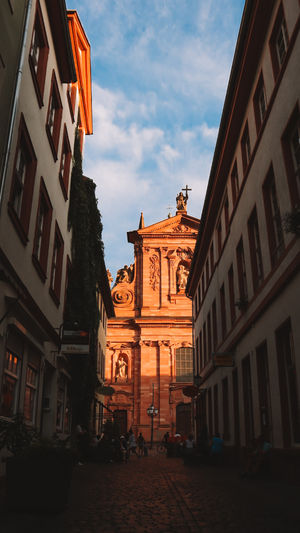 Foto von einer engen Gasse mit hohen historischen Gebäuden im Barock- oder Renaissance-Stil und einem beleuchteten Gebäude im Hintergrund, Heidelberg