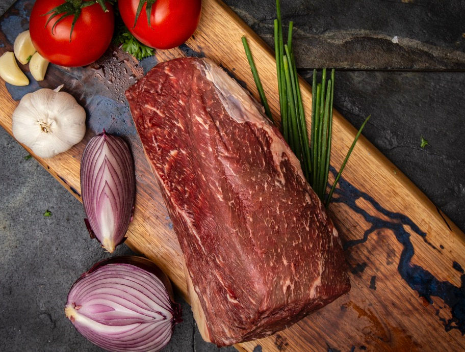 Wagyu roast on wooden cutting board with onions, garlic and tomatoes.