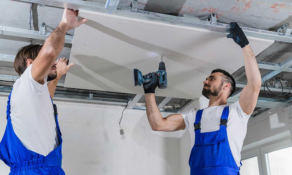 Two workers in blue overalls install a ceiling panel using a drill. They're focused, in a room with exposed beams and white walls.