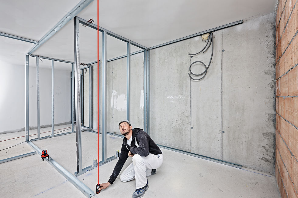 Man in construction setting measuring with laser level, framing walls. Background: concrete and exposed brick walls. Focused expression.