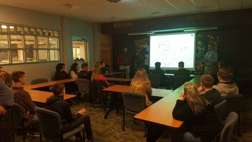 Presenter stands next to projection screen while discussing engineering program with students who are seated at tables