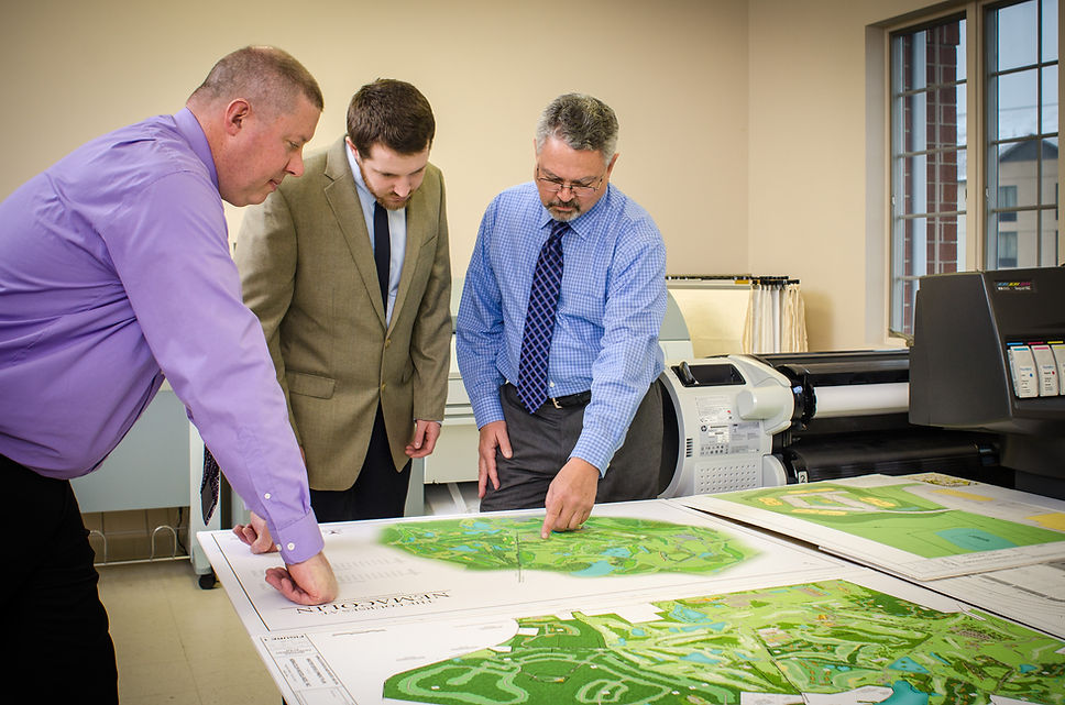 Three engineering professionals standing at table while pointing to and discussing renderings