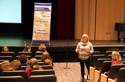 Female presenter speaking to large audience in auditorium with presentation being projected on stage