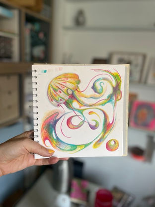 Hand holding a sketchbook showing a colourful abstract rainbow swirl jellyfish drawing in a softly blurred room.