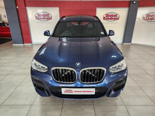 Blue BMW SUV in a showroom with red and white walls, labeled "Global Autohaus." Sleek design and shiny finish.