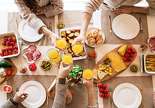 Pessoas brindando numa mesa com refeições. Aparece somente mãos em braços de quatro pessoas. A foto foi tirada de cima. Na mesa contem alimentos variádos. Abacate,tomate,pimemta vermelha,azeitona,queijo fatiado,milho cozido,pão fatiado, sadada de verduras, pratos brancos, garfos e facas. As pessoas brindam com suco de laranja nos copos.