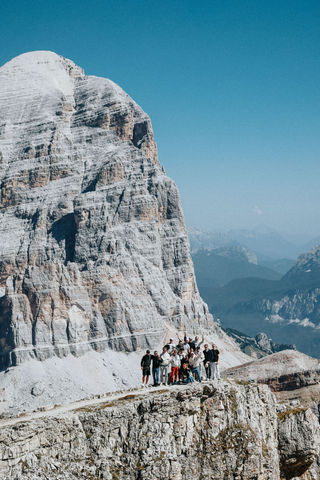Eine Gruppe von glücklichen Menschen steht auf einem Gipfel und winkt dem Fotografen. Im Hintergrund ist ein großer felsiger Berg zu sehen.