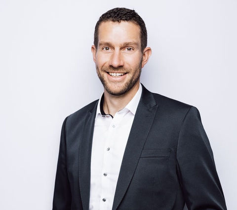 Ein junger Mann steht locker mit der linken Hand in der Hosentasche vor einem weiß-grauen Hintergrund und lächelt in die Kamera. Er trägt ein schwarzes Jacket und ein weißes Hemd