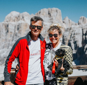 Ein gut gelauntes Paar lächelt in die Kamera, rechts steht eine Frau mit einem kleinen Hund auf dem Arm, daneben ein Herr mit Sonnenbrille und einer roten Jacke.