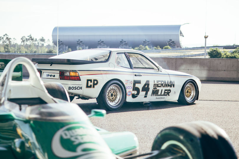 Im Vordergrund ist ein grünes Rennauto zu sehen, dahinter steht ein weißer Porsche mit Rennsport Beschriftung. Im Hintergrund ist eine große Tribühne des Hockenheimrings zu sehen. 