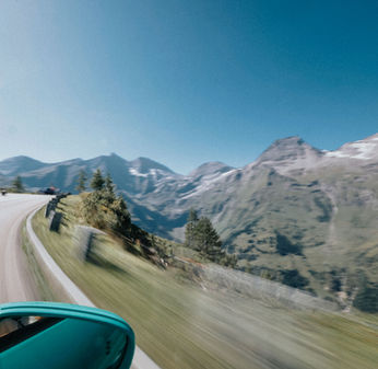 das Bild zeigt die Straße der Großglockner Hochalpenstraße aus der Perspektive eines Beifahrers. Im Hintergrund sind hohe Berge mit kleinen Schneefeldern zu sehen.