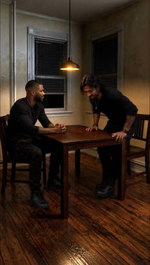 Two men lean toward each other across a wooden table in a dimly lit room.
