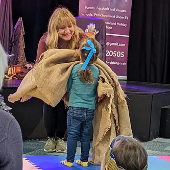 Amanda Kane-Smith is smiling as she leans forward and hands a big piece of material to a young girl during a performance of Fred the Cat's Magical Monthly Story club.jpg