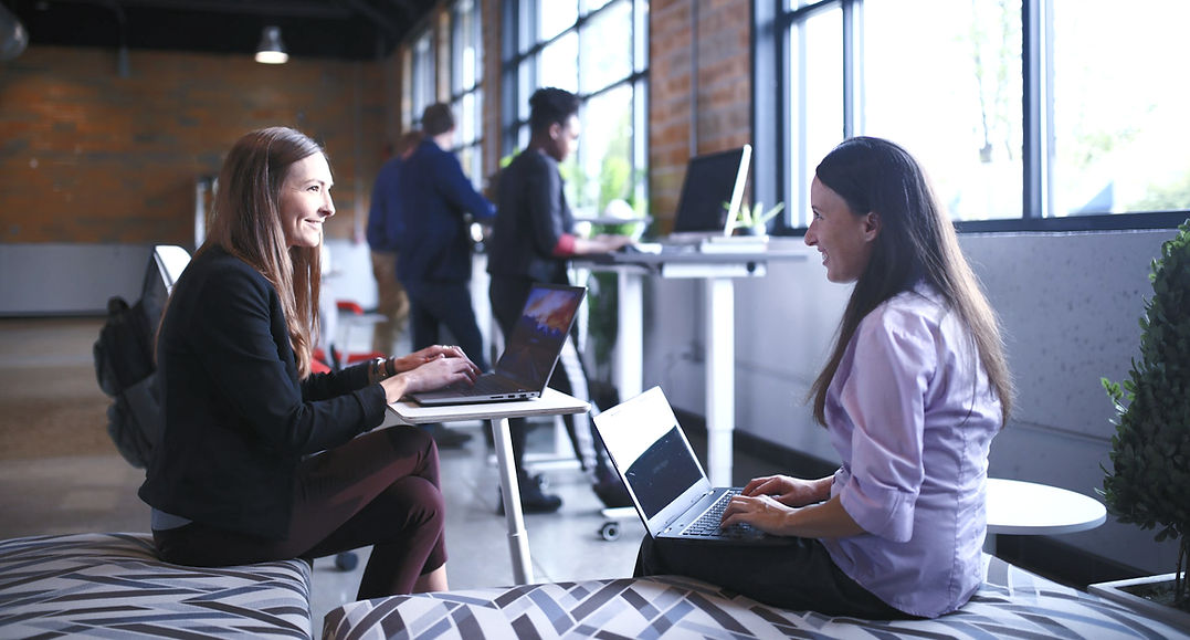 alt=" employees in a workplace chatting and working on their laptops"