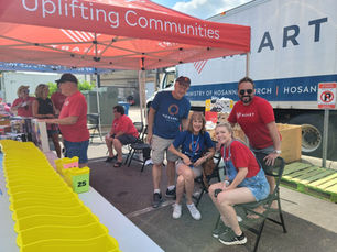 Volunteers from HEART and Hosanna Church sit under the HEART canopy smiling while others hand out drawing tickets