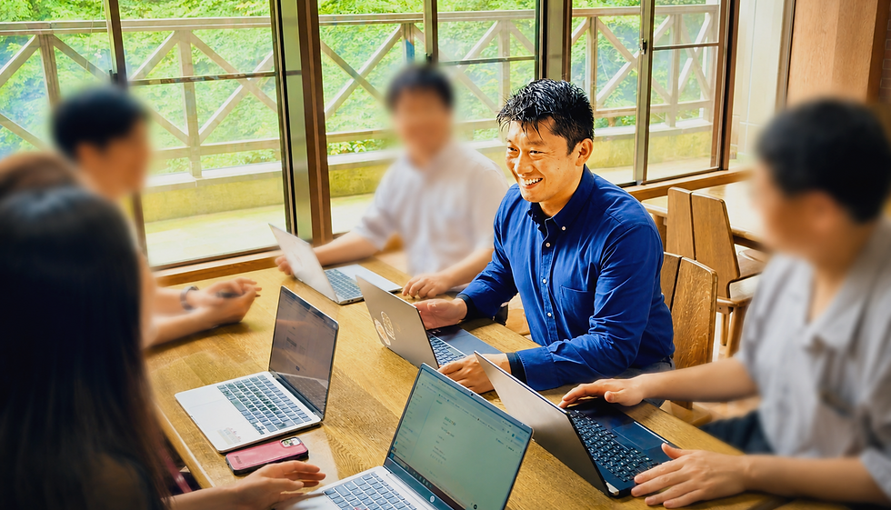 名古屋投資クラブの投資勉強会の様子。レンガ調の壁の前で、グレーのジャケットを着た男性が笑顔で話し、テーブルを囲む参加者が耳を傾けている。机の上にはノートとペットボトルがある。