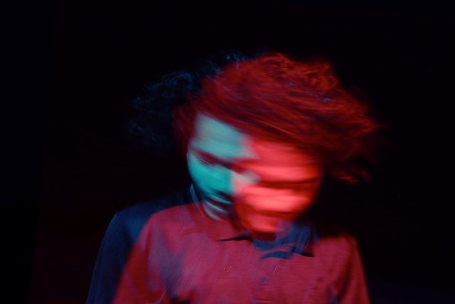 Blurry image of person appearing to shake their head from side to side, but with the effect that suggests they are in an altered state and not fully conscious. The colors blue and red in the head contrast to a black background.