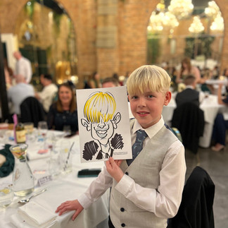 A woman in a pink dress smiles, holding a caricature of herself. She's in a brick-walled room with chandeliers and holds a glass. dalton old pump house durham