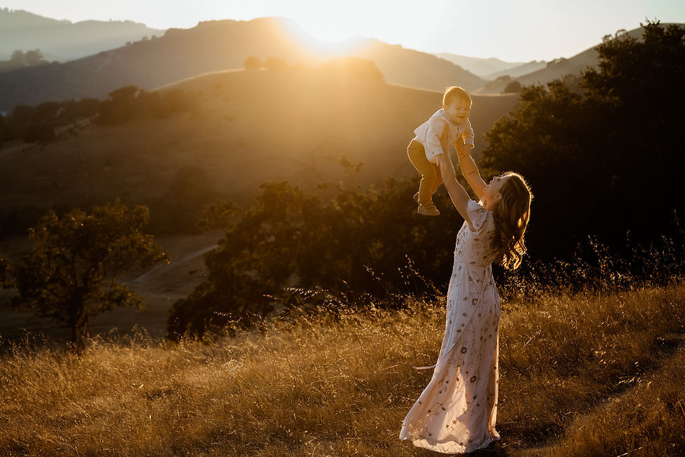 Woman in a floral dress lifts a smiling child under a golden sunset in a serene field with hills in the background. Warm and joyful mood.
