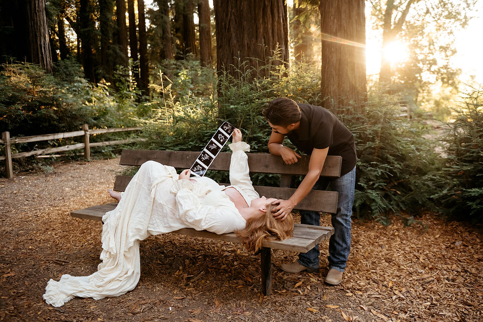 Man gently touches woman lying on a bench holding ultrasound photos. Sunlit forest backdrop with trees and wooden fence.