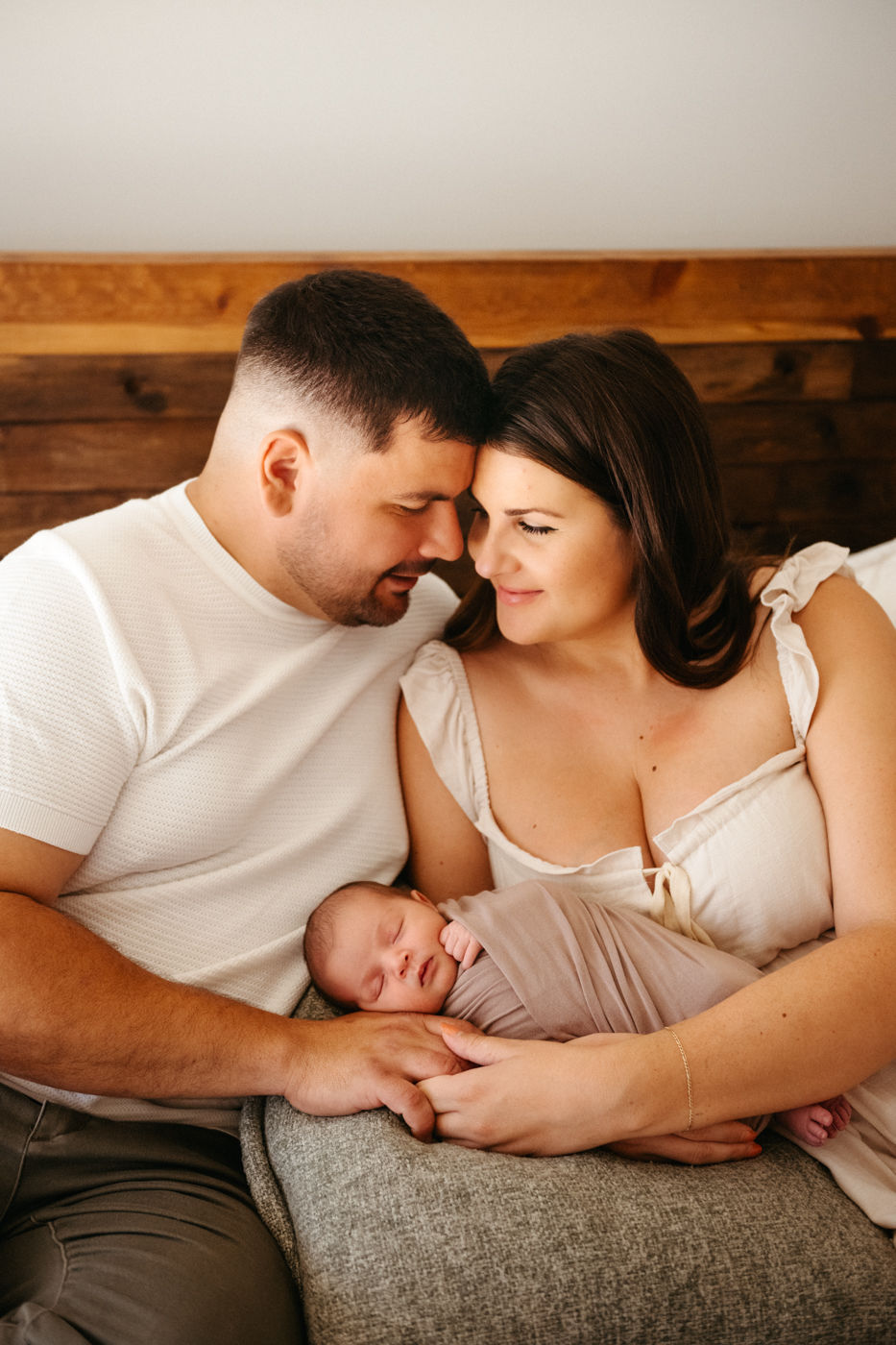 A couple lovingly holds a sleeping baby wrapped in a soft beige blanket. They're seated on a couch with a warm wooden backdrop.