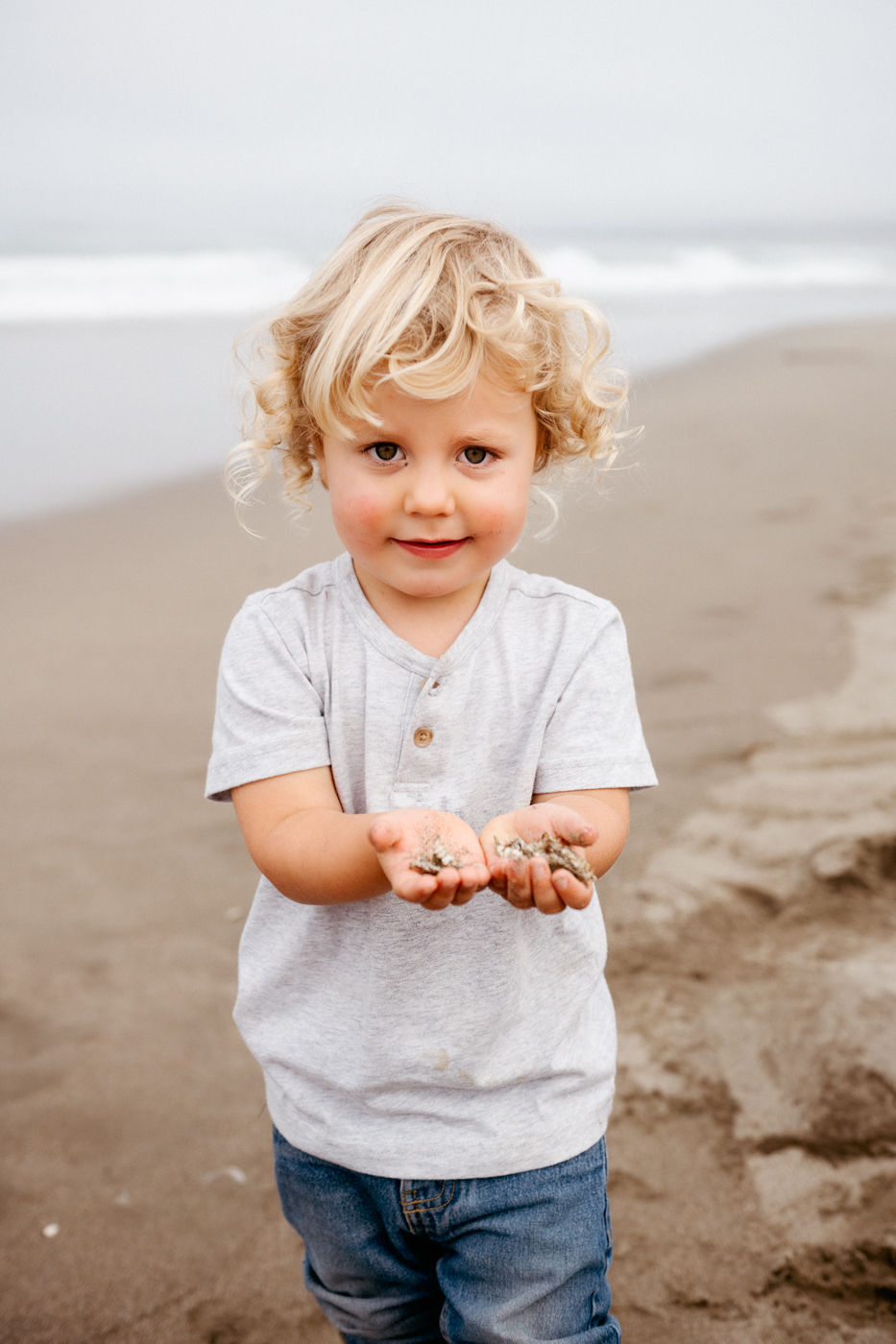 Young child with curly blonde hair holds sand in both hands, standing on a cloudy beach. Wears grey shirt and jeans, appears happy.