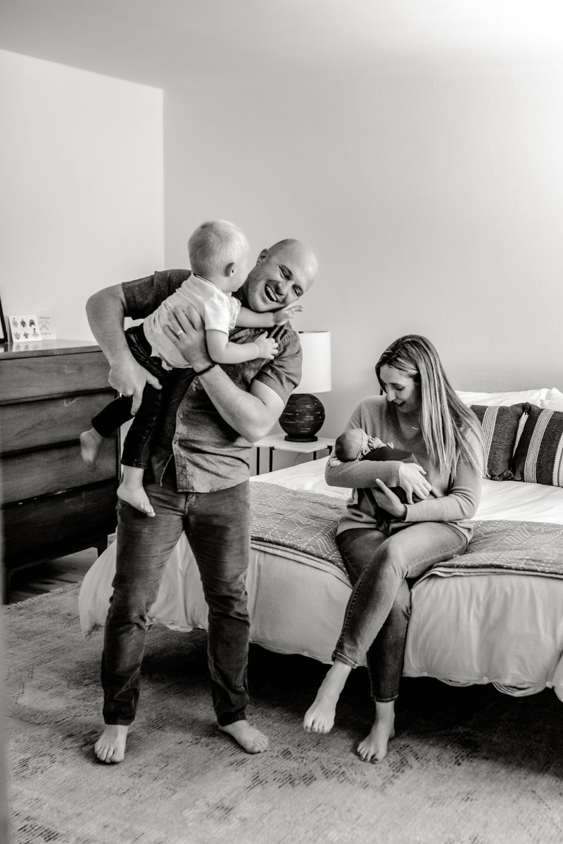 Black and whitle photo of a dad playing with his son and tossing him on the bed, while mom is in the background lovingly looking down and holding their baby girl.