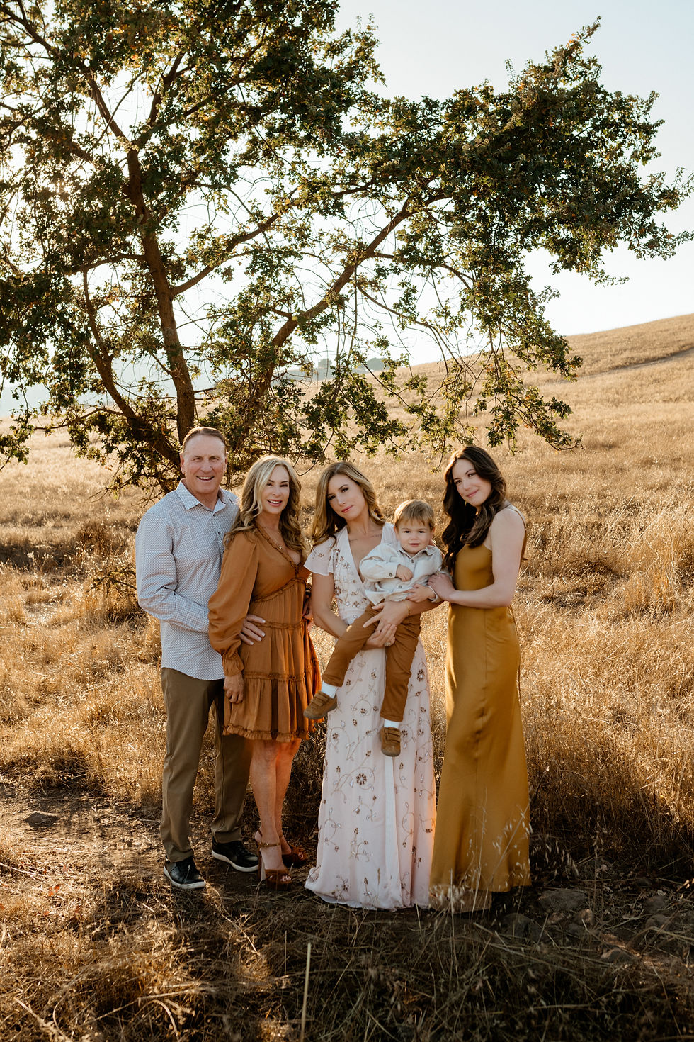 Family of five poses in a sunny field with a tree. They're wearing earth-toned outfits and smiling, creating a warm, cheerful mood.