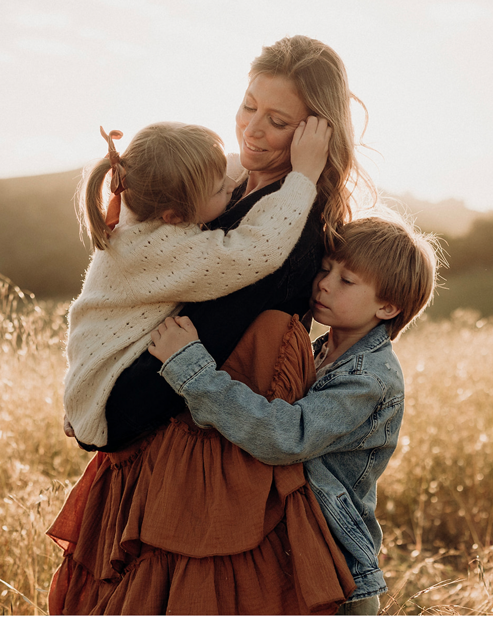 A woman lovingly embraces two children in a sunlit field. One child hugs her neck, the other holds her waist. Warm, golden tones. a beautiful photograph of Diana Jex photography, a great and affordable family and maternity photographer serving petaluma.