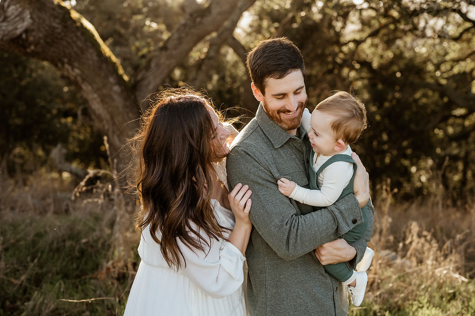 A smiling family with a man holding a baby and a woman embracing them, standing outdoors with trees in the background during sunset.