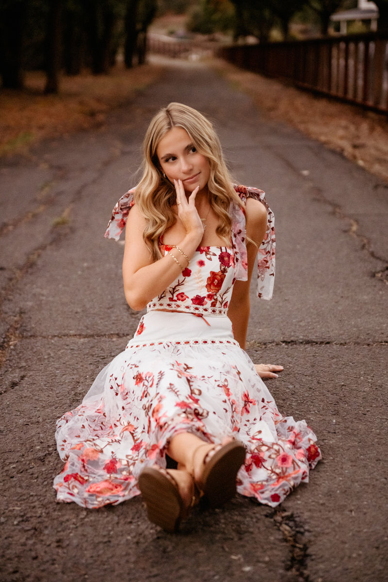 Woman in floral dress sits on a road, leaning on hand with thoughtful expression. Trees and railings line the background, creating a serene mood.