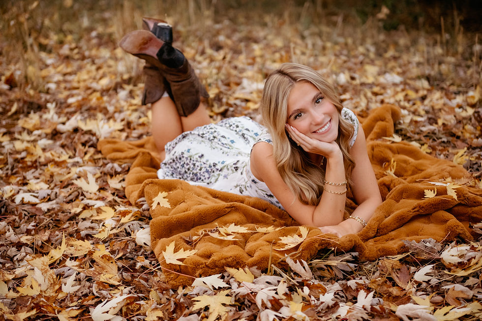 Woman in white floral dress lies on blanket in a field of autumn leaves, smiling with hands on her face. She wears brown boots, creating a cozy, joyful vibe.