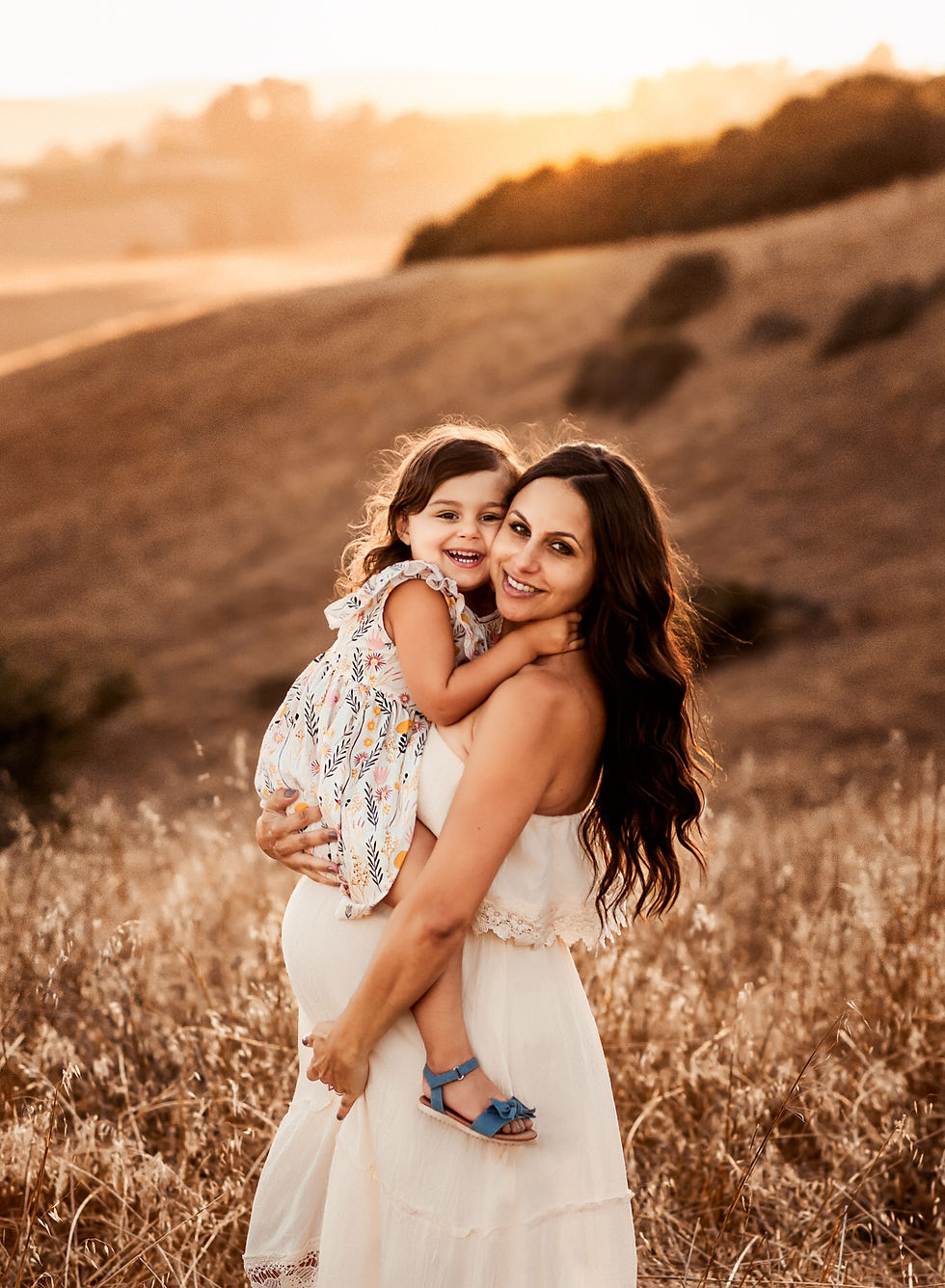 Woman in white dress holding a smiling child in a floral dress. Golden sunset over grassy hills in the background, creating a warm ambiance.