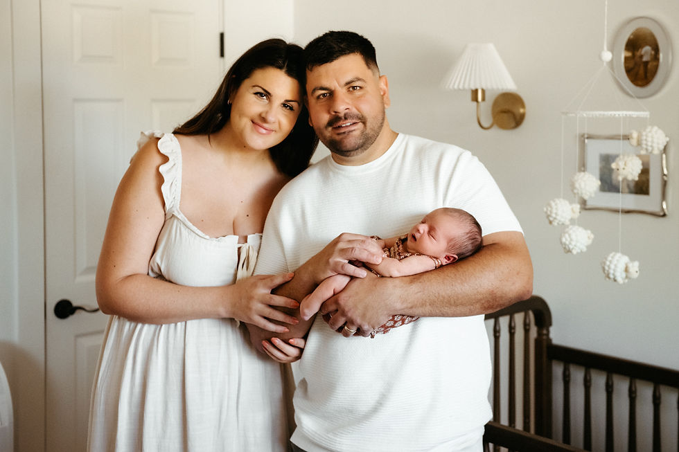 A couple smiles at the camera, holding a sleeping newborn in a cozy nursery. Neutral tones, soft lighting, and a mobile in the background.