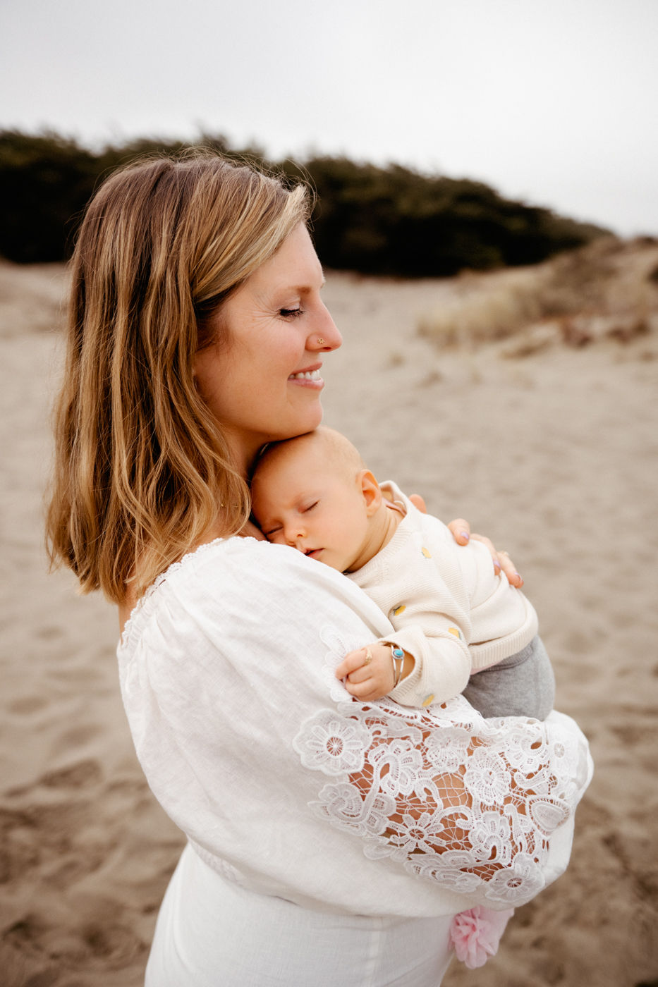 Woman smiling, holding a sleeping baby on a sandy beach. She's wearing a white dress with lace. The sky is cloudy, creating a peaceful mood.